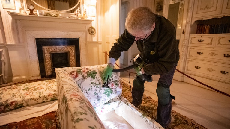 A man vacuums under the cushions of a chintz-covered sofa